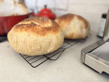 Two golden, crusty loaves of freshly baked bread cool on wire racks in a bright kitchen, accompanied by kitchen appliances and a red Dutch oven.