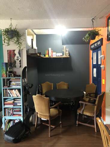 L'image représente une pièce confortable avec une table ronde et des chaises, une bibliothèque et des œuvres d'art, avec une porte de boîte de police et des plantes, dans une atmosphère chaleureuse et accueillante.
