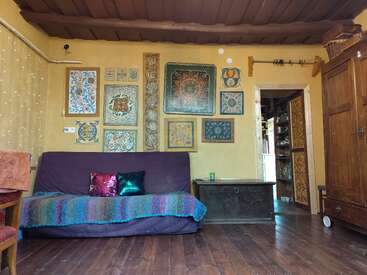 In einem gemütlichen Zimmer mit gelben Wänden und Holzböden gibt es bunt gemusterte Kunst, eine lila Couch mit bunten Kissen, eine Truhe und einen rustikalen Holzschrank.