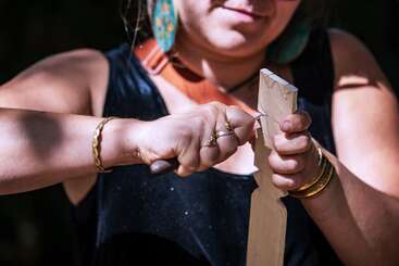 Eine Frau schnitzt ein Stück Holz mit einem kleinen Messer. Sie trägt Goldschmuck, ein schwarzes ärmelloses Oberteil und große türkisfarbene Ohrringe und konzentriert sich.