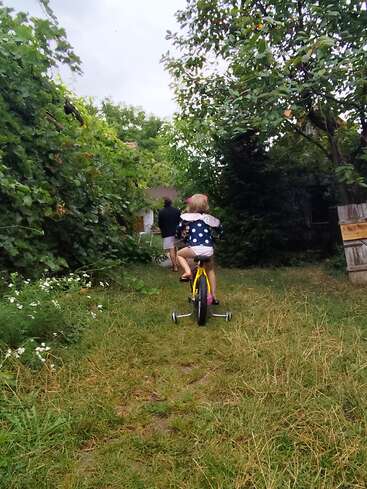 Ein Kind fährt auf einem gelben Fahrrad mit Stützrädern einen grasbewachsenen Gartenweg entlang, gefolgt von einer Person, umgeben von üppig grünem Laub und Bäumen.