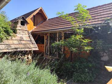 Ein rustikales Haus mit rotem Ziegeldach, Holz- und Backsteinelementen, üppigen Grünpflanzen, Lavendel, einem Steinofen und einem klaren blauen Himmel an einem sonnigen Tag.