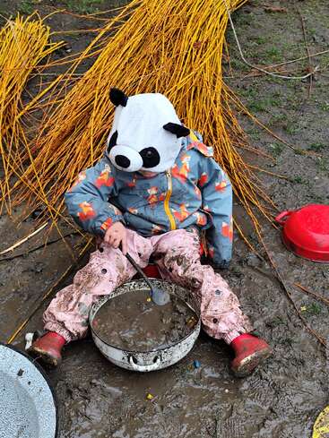 A child wearing a panda hat and fox-patterned raincoat sits in mud, stirring a pot with a spoon, surrounded by yellow sticks and muddy ground.