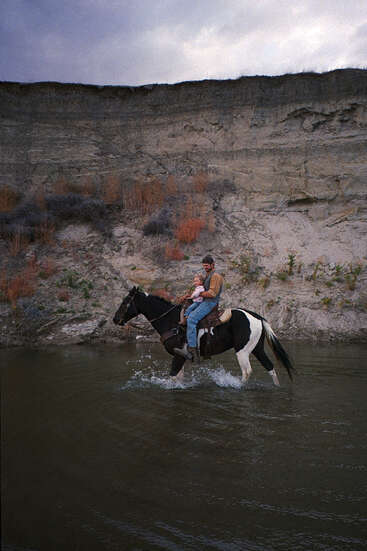 Un hombre y un niño montan un caballo blanco y negro por aguas poco profundas bajo un acantilado rocoso. El cielo está nublado, lo que añade una atmósfera melancólica.