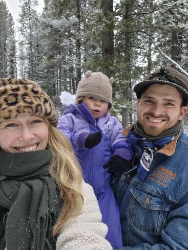Una familia sonriente posa al aire libre en un bosque nevado. La mujer lleva un sombrero con estampado de leopardo, el hombre una gorra y el niño va abrigado con ropa morada.