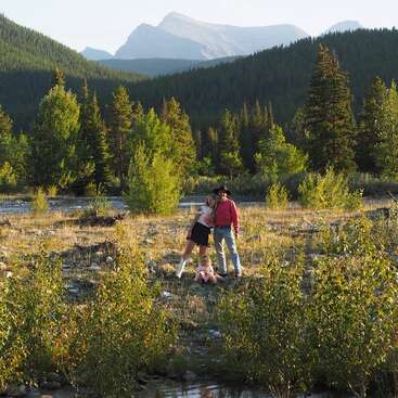 A family stands together in a scenic, grassy field surrounded by green trees. Majestic mountains rise in the background under a clear blue sky.