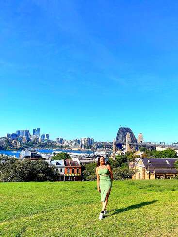Eine Frau in einem grünen Kleid steht auf einem grasbewachsenen Hügel, mit der Sydney Harbour Bridge, der Skyline der Stadt und dem blauen Himmel im hellen, sonnigen Hintergrund.