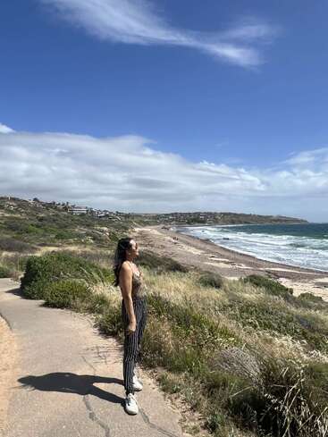 A woman stands on a coastal path, basking in the sun. Behind her, the ocean waves crash gently onto the shore under a bright, blue sky.