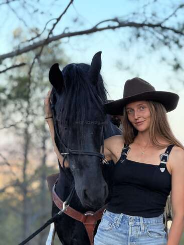Une jeune femme coiffée d'un chapeau à large bord se tient à côté d'un cheval noir, lui tenant doucement la tête. Les arbres se fondent dans le flou à l'arrière-plan. Elle sourit doucement, profitant de la nature.