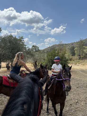 Une femme et un enfant, tous deux à cheval, traversent une campagne pittoresque. Le ciel est partiellement nuageux, et les montagnes et les arbres les entourent magnifiquement.