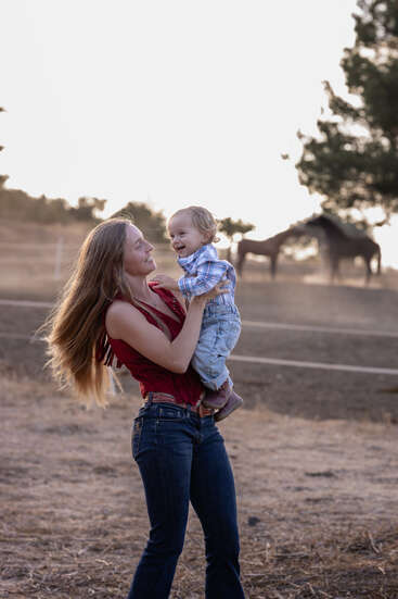 Une femme soulève joyeusement un enfant souriant à l'extérieur d'un ranch. Des chevaux paissent en arrière-plan. Le soleil couchant diffuse une lumière chaude et sereine.