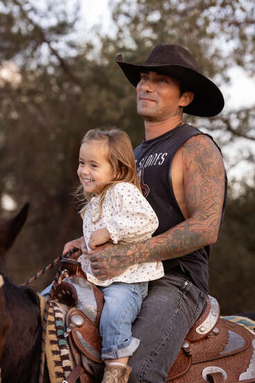 Un homme tatoué coiffé d'un chapeau de cow-boy tient une jeune fille souriante sur ses genoux tandis qu'il chevauche un cheval. Ils semblent heureux, profitant d'un moment paisible en plein air.