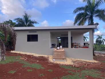 This image shows a single-story, modern gray house with a porch, surrounded by palm trees, red soil, and patches of grass under a partly cloudy sky.