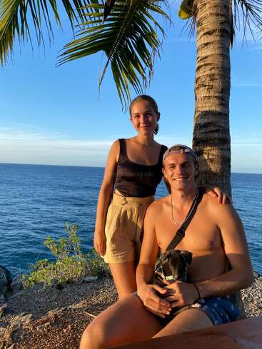 A smiling couple poses by the ocean at sunset, standing near a palm tree. The man is shirtless, sitting, holding a small black-and-white dog.