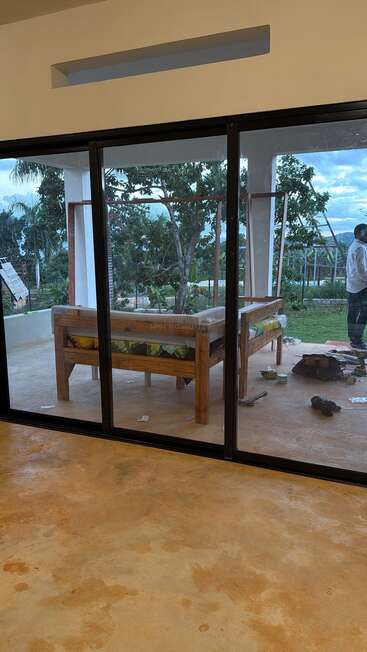 This image shows a patio viewed through large sliding glass doors. Outside, a wooden bench, tools, and a person standing near lush, green trees and mountains.