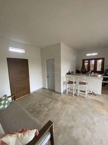 This image shows a minimalist living space with a wooden door, a small couch, a breakfast bar with two stools, and a cat near the kitchen area.
