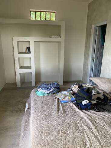 A minimalist room with concrete floor, beige bedspread, and open shelving. Clothes and books are scattered on the bed. Small window and doorway visible.