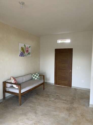 This minimalist room features a wooden sofa with cushions, a closed wooden door, a small window above, botanical wall art, and a bare lightbulb ceiling fixture.