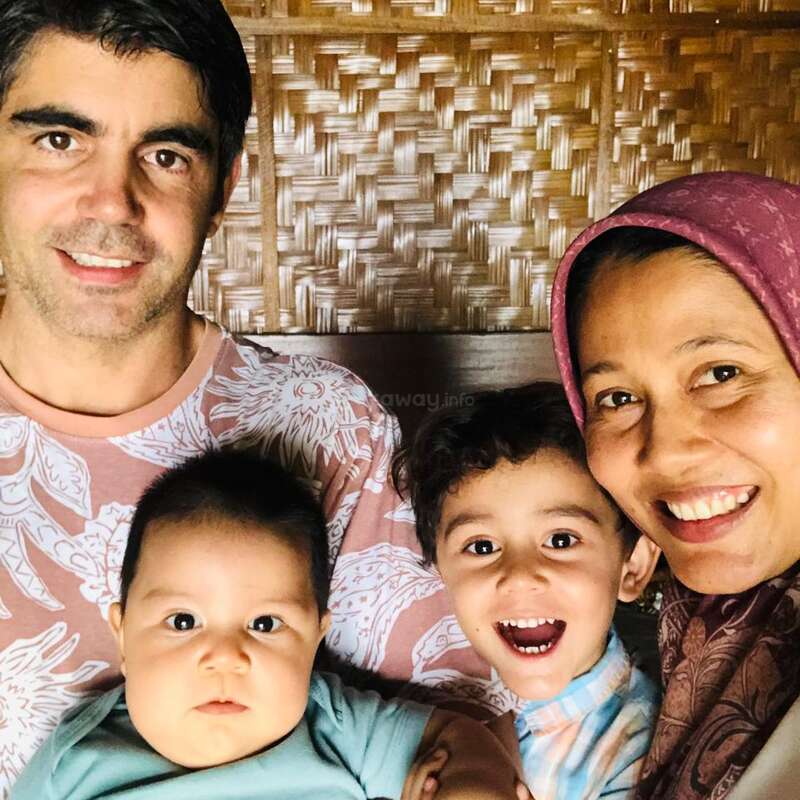 A happy family of four poses closely together, smiling warmly. The father and mother hold their two young children, capturing a joyful, loving moment indoors.