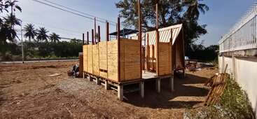 Auf einer erhöhten Plattform mit Teilwänden und senkrechten Balken wird gerade ein Holzhaus gebaut. In der Umgebung gibt es Bäume, Stromleitungen und einen klaren, sonnigen Himmel.