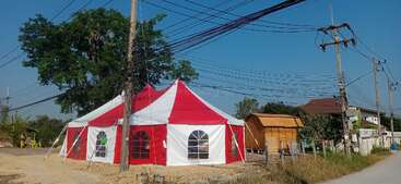 Una tienda roja y blanca se encuentra cerca de un camino de tierra, rodeada de cables eléctricos, árboles y una pequeña estructura de madera, todo ello bajo un cielo azul claro.