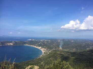 Un paysage côtier pittoresque comprenant une baie, des collines verdoyantes, des petites villes, un panache de fumée, une mer bleue et un ciel partiellement nuageux sous un soleil éclatant.