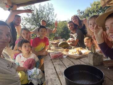 Eine Gruppe von Erwachsenen und Kindern versammelt sich um einen rustikalen Holztisch im Freien, teilt Essen und lächelt und genießt einen sonnigen Tag in einer entspannten, fröhlichen Atmosphäre.