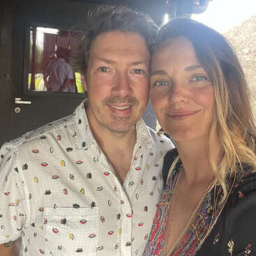 A smiling couple poses closely together for a selfie indoors. The man wears a patterned shirt, and the woman has a nose ring and loose, wavy hair.