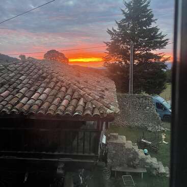 Ein rustikales Haus mit einem Ziegeldach steht unter einem dramatischen Himmel bei Sonnenuntergang. In der friedlichen Szenerie sind Bäume, Steinmauern und ein blauer Lieferwagen zu sehen.