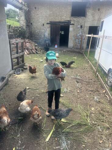 A child wearing a green hat stands outside on a farm, gently holding a chicken. Several other chickens are pecking around on the ground nearby.