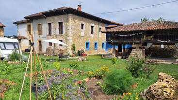 A rustic stone house with blue-trimmed windows sits beside a lush garden, caravan, and barn. Sunny yard features plants, flowers, benches, and festive bunting.