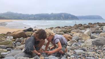 Two children crouch on a rocky beach, intently examining stones. The background features waves, sandy shore, and green hills under a cloudy sky, evoking peaceful curiosity.