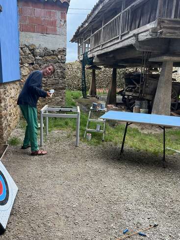 A person stands outdoors beside a stone building, painting furniture. There’s a blue target, arrows on the ground, and a rustic wooden structure in the background.