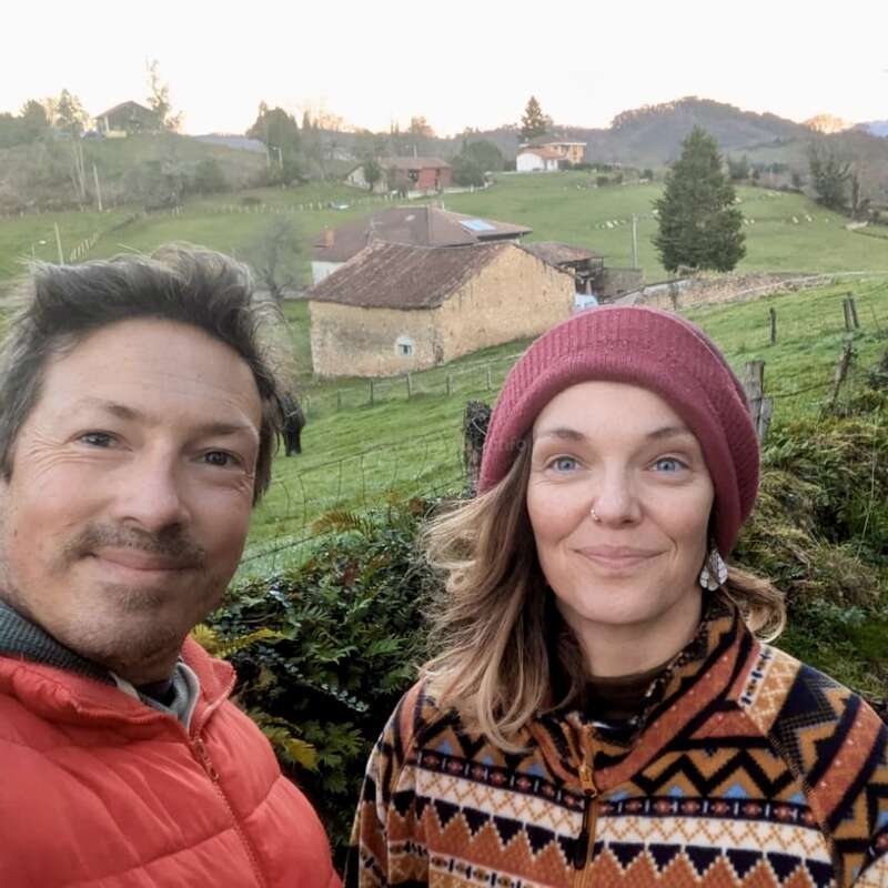 A man and woman smile outdoors in a rural setting, wearing warm clothes. Behind them are green fields, rustic houses, trees, and distant hills at sunset.