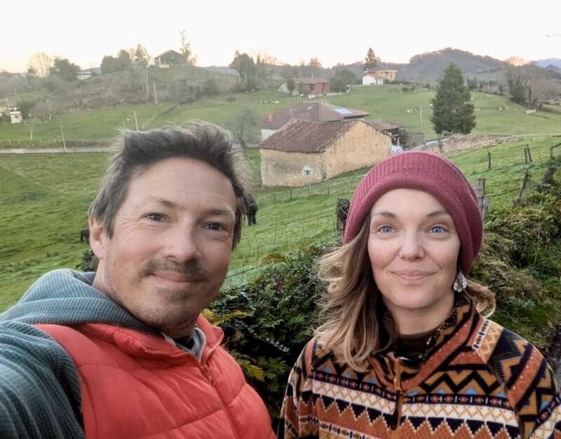 A man and woman smile outdoors in a rural setting, wearing warm clothes. Behind them are green fields, rustic houses, trees, and distant hills at sunset.