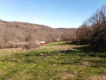 A sunny landscape features green grass, rolling hills, leafless trees, and a red round structure with a white roof under a clear blue sky. Peaceful scene.