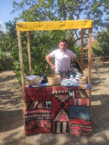 L'image représente un homme debout derrière un stand de marché en plein air, entouré d'arbres et d'un ciel bleu clair, avec un tapis coloré et des paniers.