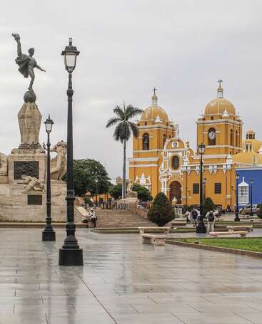 Une magnifique place abrite une église coloniale jaune à deux dômes, une statue impressionnante, des palmiers, des lampadaires, des jardins verdoyants et des gens qui profitent du paysage.