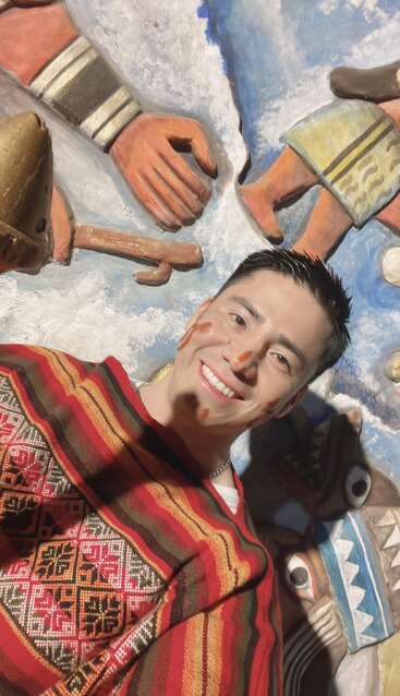 Un hombre sonriente vestido con un poncho tradicional de colores vivos con motivos rojos, amarillos y verdes. Lleva la cara pintada y posa ante un fondo artístico muy colorido.