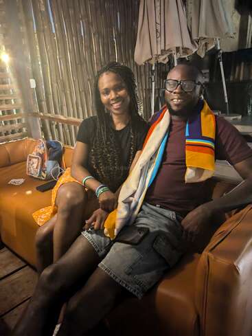 A smiling man and woman sit closely together on a brown couch in a cozy, rustic setting, looking relaxed and happy. Warm lighting enhances the atmosphere.