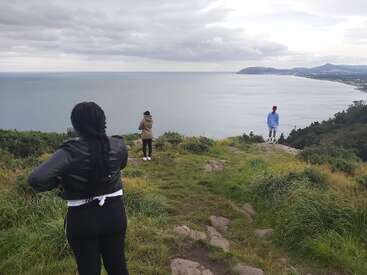 Three people stand atop a grassy cliff overlooking a vast, calm sea. Two take photos while one gazes out, with cloudy skies above and distant hills.