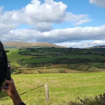 A person photographs a scenic landscape of rolling green hills, patchwork fields, distant mountains, and vibrant blue sky dotted with fluffy white clouds on a sunny day.