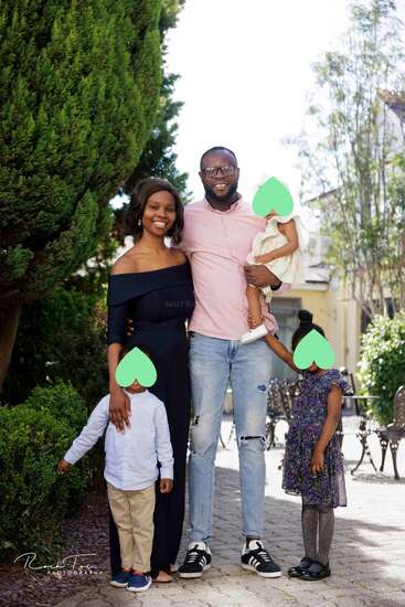 A smiling family of five poses outdoors. The parents stand in the middle, holding their three young children. The children’s faces are covered with green hearts.