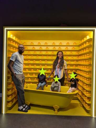 A family poses inside a bright yellow display filled with rubber ducks. The children sit in a yellow bathtub, their faces covered by green stars.