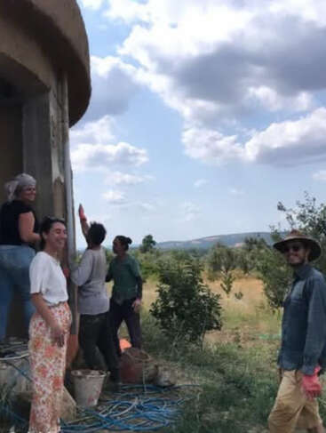 Cinco pessoas trabalham alegremente em um prédio redondo e rústico sob um céu parcialmente nublado. Baldes, mangueiras e vegetação os cercam, criando uma atmosfera descontraída e comunitária ao ar livre.