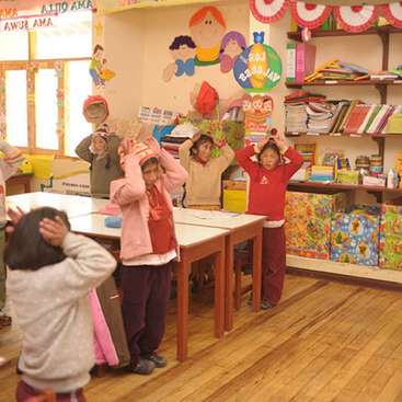 A imagem mostra uma sala de aula animada, com crianças em pé ao redor das mesas, com decorações coloridas e prateleiras de livros e materiais adornando as paredes e o chão.