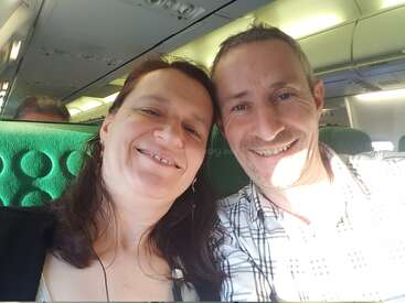 Ein lächelndes Paar macht ein Selfie zusammen, während es in einem Flugzeug sitzt. Das Sonnenlicht beleuchtet ihre Gesichter, und hinter ihnen sind grüne Sitze zu sehen.