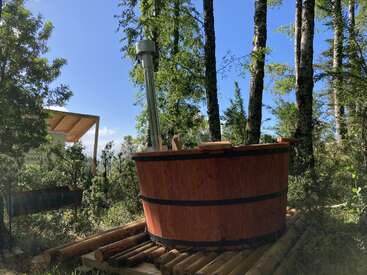 Una bañera de madera con una chimenea metálica se encuentra en un frondoso bosque verde, rodeada de árboles y luz natural, creando un refugio tranquilo.