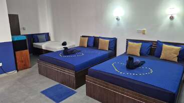 Esta imagem mostra um quarto de hotel limpo e moderno com duas camas de casal e uma cama de solteiro, roupa de cama azul, cisnes decorativos na toalha, travesseiros amarelos e luzes de parede.