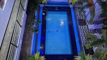 Esta imagem mostra uma piscina retangular iluminada cercada por azulejos azuis e palmeiras, com espreguiçadeiras em um deck estampado. A foto é tirada de cima.